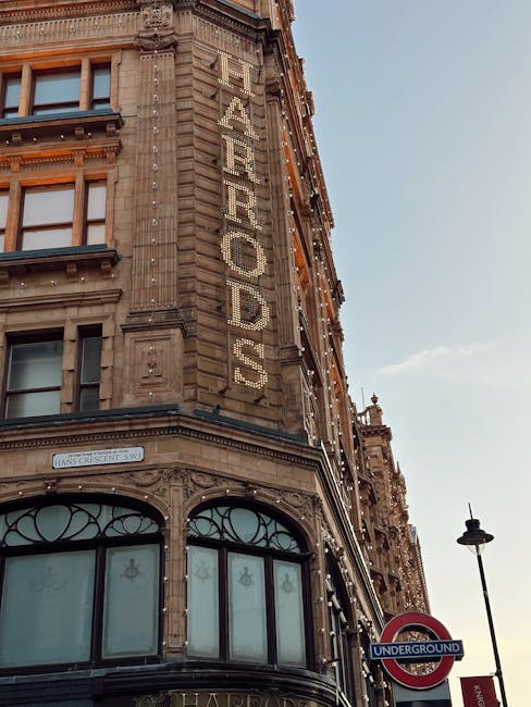Exterior view of a historic building on Brompton Road near Harrods, displaying a large vertical sign with the word 'FEARRODS' made of illuminated bulbs. The building features ornate architectural details with stone carvings and large, curved windows with decorative metalwork. The scene is captured during daylight with a clear sky, and a black lamppost and an underground station sign are visible in the foreground. Brompton Carpet Cleaning specializes in deep cleaning and surface sanitisation of residential and commercial interiors, ensuring thorough hygiene and maintained appearance, reflecting their expertise in domestic cleaning services.
