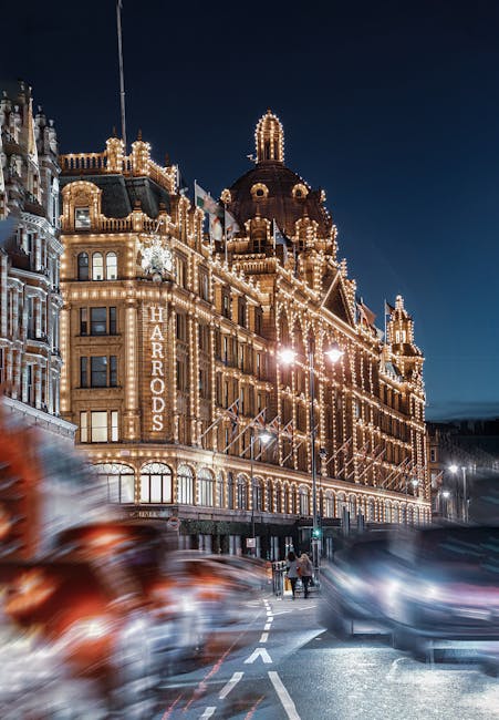 A nighttime view of the historic Harrods building illuminated with numerous decorative string lights, highlighting its ornate architecture with domed rooftops and intricate detailing. The building is situated along Brompton Road, with blurred motion of cars on the street in front, and pedestrians walking nearby. The scene is set against a dark sky, showcasing the vibrant glow of the facade lighting. This image relates to the context of domestic and commercial cleaning services by emphasizing the importance of maintaining cleanliness and hygiene in prominent, high-traffic locations, as promoted by Brompton Carpet Cleaning for properties near Harrods.
