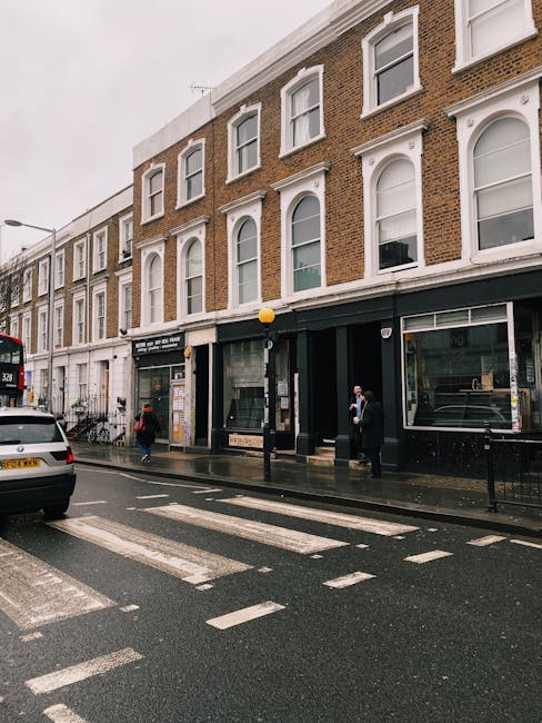 Street view of a three-story brick building on Old Brompton Road near South Kensington, featuring large ground-floor windows with black framing, a black entrance door, and a black storefront awning. The pavement is wet, indicating recent rain, with a pedestrian crossing in the foreground and a few people walking or standing on the sidewalk. A white car is parked nearby, and the overcast sky creates a subdued lighting atmosphere. Brompton Carpet Cleaning is visible on the website, emphasizing surface cleaning and deep cleaning services in the local area.