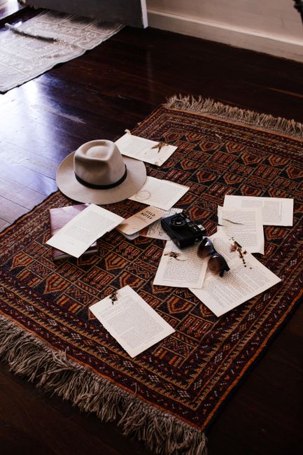 A close-up view of a richly patterned, dark-colored Persian rug on a polished wooden floor in a living room. The rug has intricate geometric and floral designs with fringed edges. Scattered across the rug are several open books, a gray felt fedora hat, a pair of black sunglasses, a black camera, and some loose papers or notes. The room is illuminated with natural light coming from the left, highlighting the clean and well-maintained appearance of the surface. Brompton Carpet Cleaning specializes in surface cleaning and deep cleaning services, ensuring hygiene and stain removal for residential spaces like Brompton flats. This scene captures a cozy, lived-in atmosphere with meticulous attention to cleanliness and maintenance.