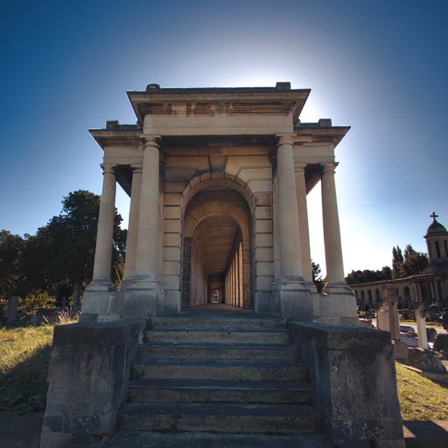 A historical stone monument featuring a rectangular entranceway supported by four large, classical-style columns with a detailed entablature on top. The monument is situated outdoors on a raised platform accessed by a short flight of stone steps, with a pathway leading through the arch into a covered corridor that extends into the background. The area is illuminated by bright sunlight, casting shadows on the stone surfaces. Surrounding greenery and other classical structures are visible in the background, emphasizing the monument's grandeur and well-maintained appearance, consistent with surface cleaning and preservation in a public or memorial setting.