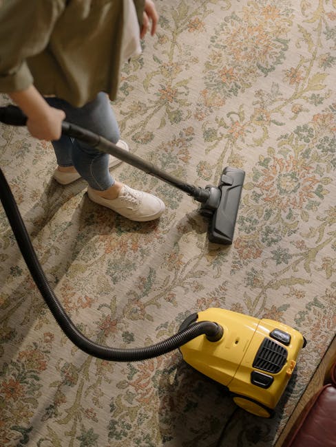 A close-up view of a richly patterned, dark-colored Persian rug on a polished wooden floor in a living room. The rug has intricate geometric and floral designs with fringed edges. Scattered across the rug are several open books, a gray felt fedora hat, a pair of black sunglasses, a black camera, and some loose papers or notes. The room is illuminated with natural light coming from the left, highlighting the clean and well-maintained appearance of the surface. Brompton Carpet Cleaning specializes in surface cleaning and deep cleaning services, ensuring hygiene and stain removal for residential spaces like Brompton flats. This scene captures a cozy, lived-in atmosphere with meticulous attention to cleanliness and maintenance.